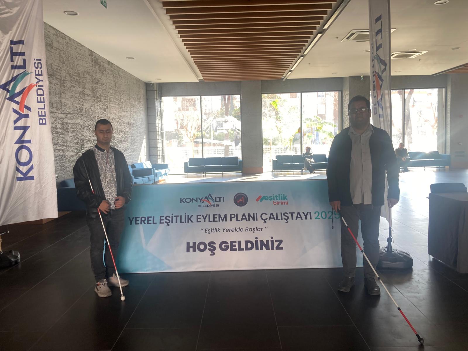 Oğuz ÖZdEMİR ve Mert YAKIŞTIR, çalıştay alanının girişinde duruyor. Her ikisi de beyaz baston kullanıyor. Arka planda, "KONYAALTI BELEDİYESİ" yazılı bir afiş ve "YEREL EŞİTLİK EYLEM PLANI ÇALIŞTAYI 2025" yazılı bir pankart bulunuyor. Pankartta ayrıca "Eşitlik Yerelde Başlar" ve "HOŞ GELDİNİZ" ifadeleri yer alıyor. Mekan modern bir tasarıma sahip, geniş cam pencerelerden dışarısı görünüyor ve içeride mavi renkli koltuklar var. Ortam aydınlık ve ferah görünüyor.